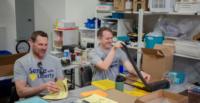 two men sit at a table and cut paper and assemble goal setting booklets